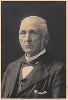 Black-and-white photograph of Joel Bean Bassett in his seventies. He is wearing a three-piece suit and bowtie and frowning, looking past the photographer to the left side of the image. 